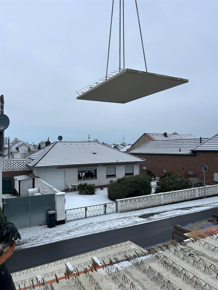 Filigrandeckenelement hängt an den Kranketten über der Baustelle, im Hintergrund verschneite Wohnhäuser in Mechernich