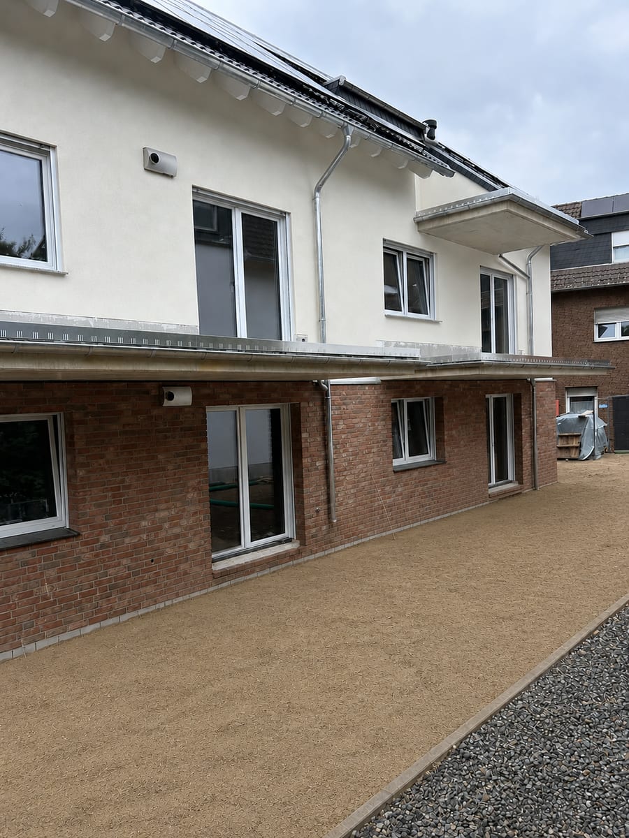 Rückseite eines fertiggestellten Wohnhauses mit Klinkersockel, verputztem Obergeschoss und Vordach, davor die profilierte Terrassenfläche mit Randeinfassung und Kiesstreifen